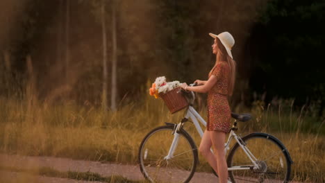 Slow-motion:-Young-sexy-smiling-blonde-woman-in-hat-and-light-brown-dress-walking-with-bike-and-flowers-in-basket-on-field-in-summer-at-sunset.-Moves-in-the-direction-of-the-camera.